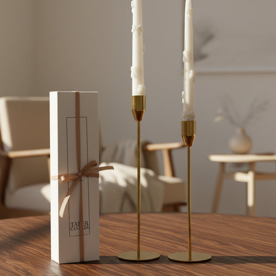 Gold candle holders with white candles on a wooden table, next to a white box with a brown ribbon.