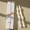 Two white candles with decorative tops next to their packaging on a beige background