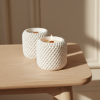 Two textured white candle holders on a wooden surface with a softly lit background.
